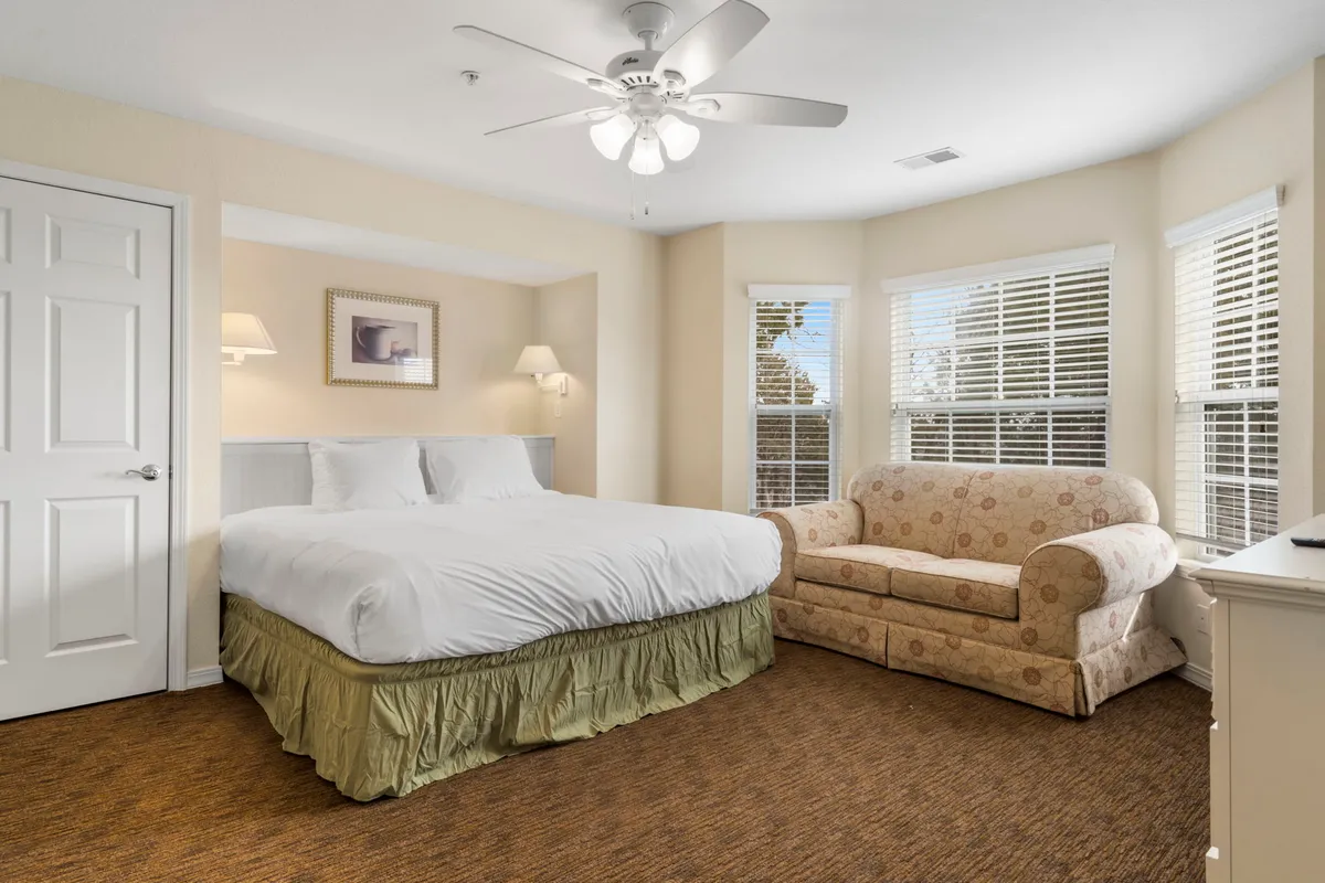 A bright, serene king bedroom with a plush bed, a cozy sofa, and sunlight streaming through large windows. The scene exudes relaxation and comfort, with subtle, thoughtful touches visible throughout the room, such as blackout shades and soft bedding.