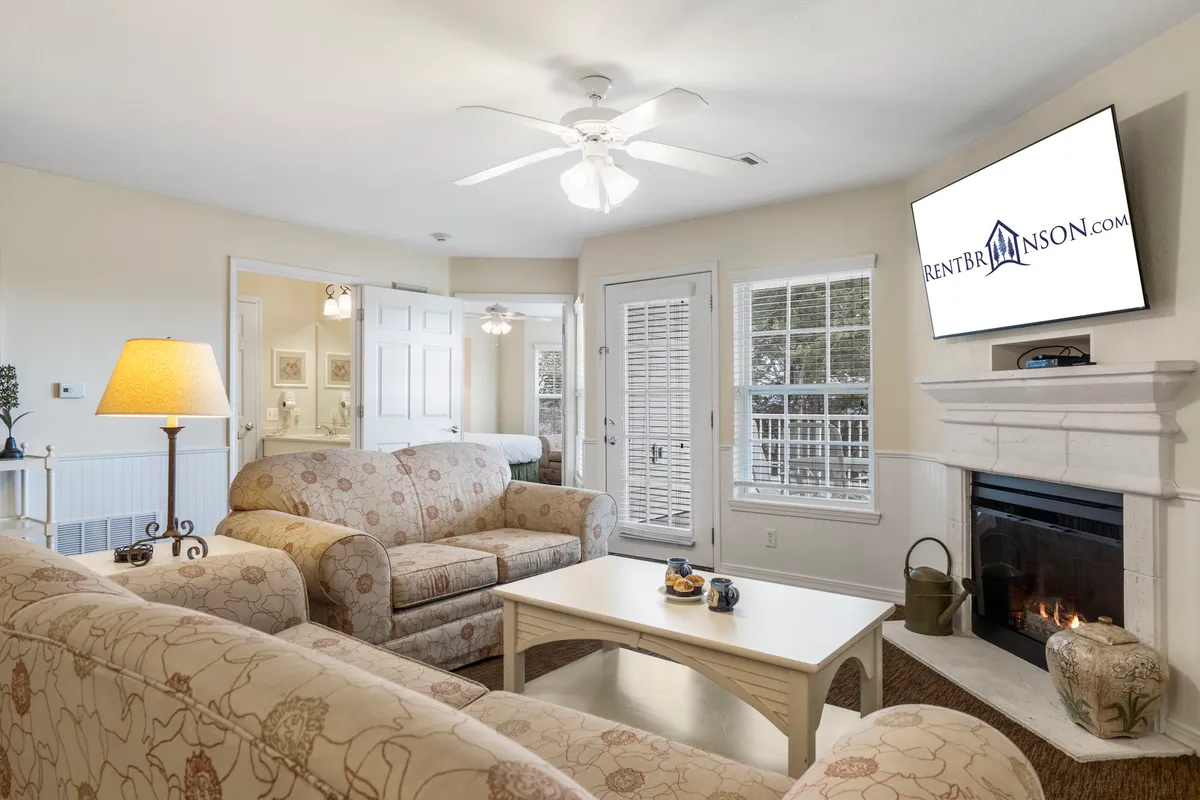 A welcoming living room featuring comfortable sofas arranged around an electric fireplace, with a television and a decorative lamp adding to the cozy ambiance. Large windows let in abundant natural light, and the room is styled to encourage relaxing, entertaining, or gathering for a movie night.