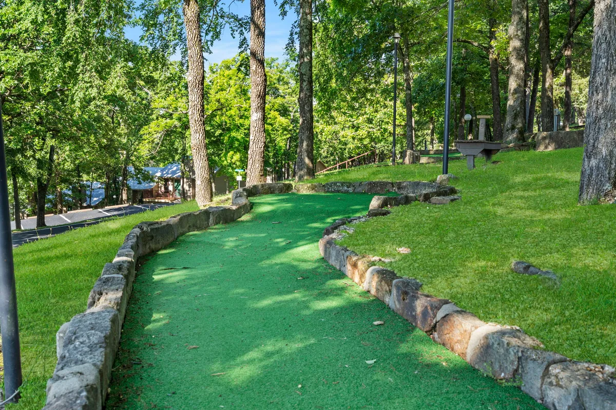 A playful, well-maintained mini-golf pathway winds through lush greenery and stone borders, inviting families to enjoy friendly competition and fun moments outdoors. The area is surrounded by trees, creating a natural and relaxing backdrop.