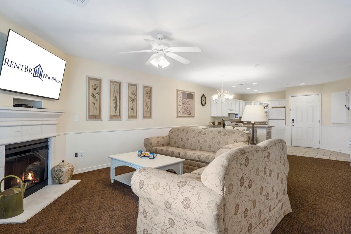 A spacious, open-plan interior with a plush sofa arranged near an electric fireplace, a television, and a dining area in the background. The fireplace anchors the room as a central gathering spot, while the open layout encourages easy conversation and a seamless flow between lounging and dining areas.