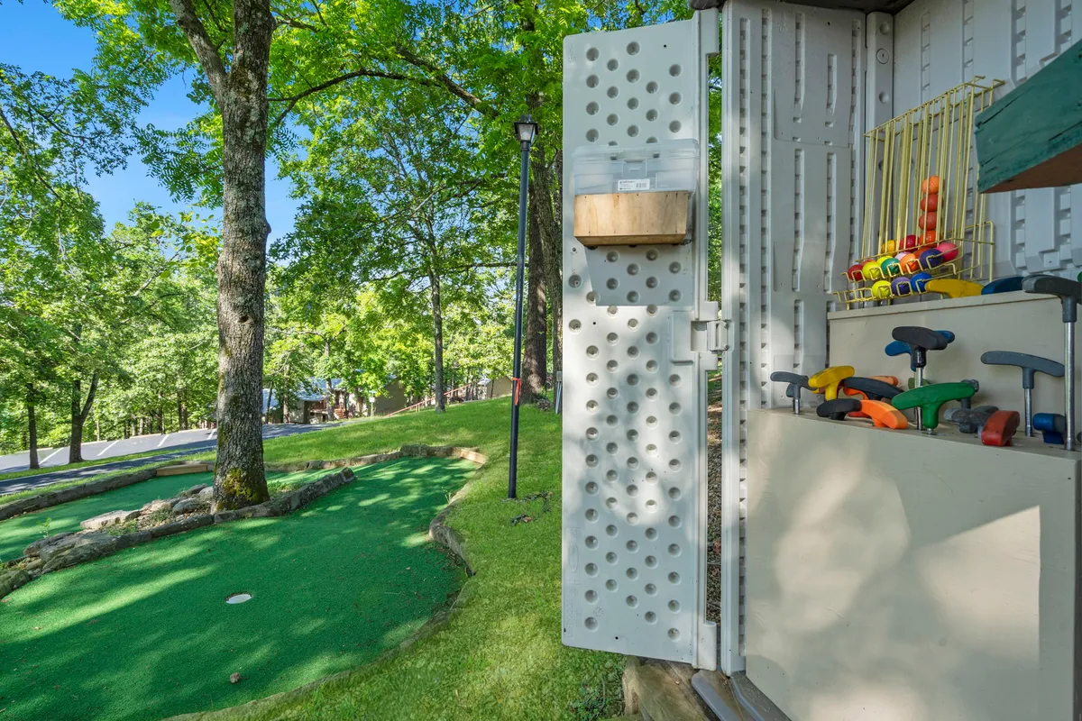 An inviting mini-golf putting green surrounded by mature trees, with a handy storage cabinet in view. The playful mood hints at family fun and friendly competition.