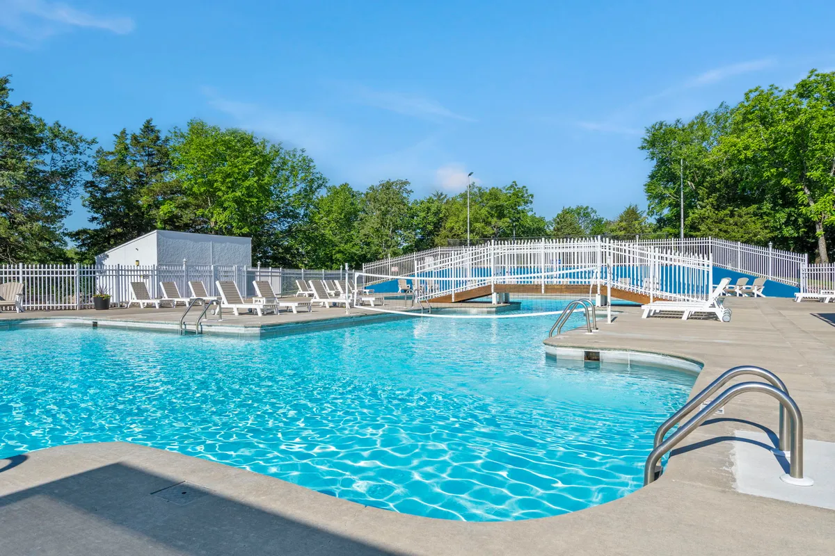 A vibrant exterior scene of the resort’s swimming pool, surrounded by lounge chairs and lush greenery. A wooden bridge crosses one end, and the atmosphere is bright and cheerful, suggesting a welcoming spot for swimming, sunbathing, or socializing.