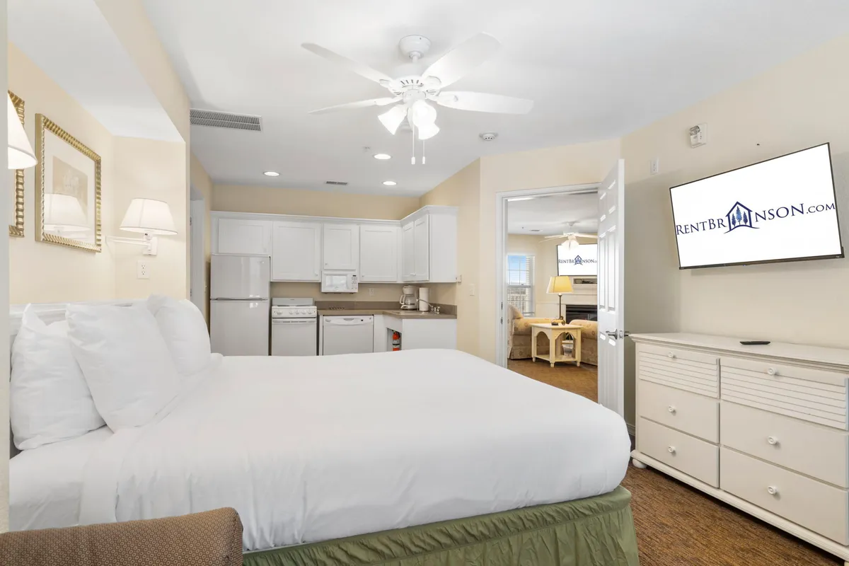 A cozy private suite featuring a comfortable bed with white linens and a compact kitchenette area. The kitchenette is visible, equipped for simple meals or snacks, while a TV on the wall suggests comfort and flexibility.