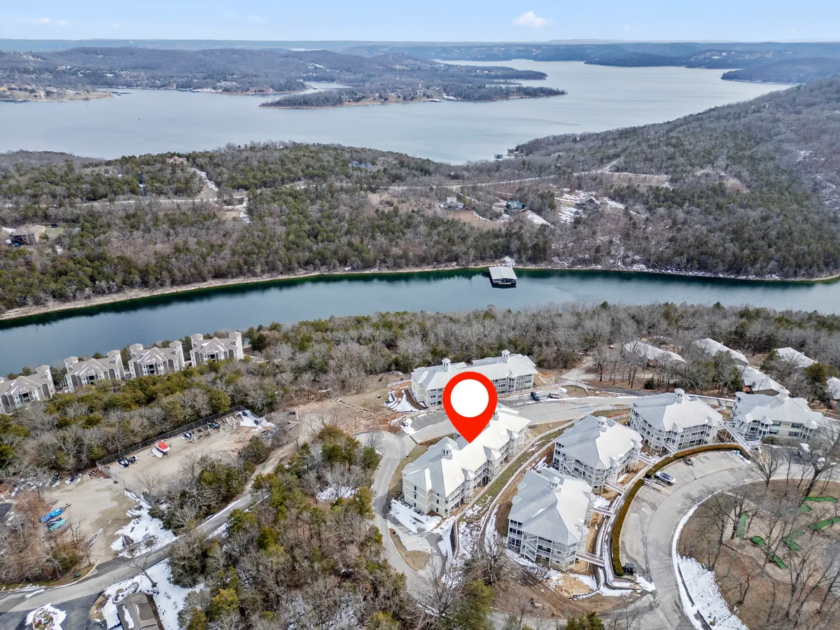 This exterior shot looks over the lake bordered by forests, with residential buildings nestled between the trees and a dock extending into the water. The composition includes both the natural beauty of the area and the human connection to it, suggesting activities such as hiking, exploring nature trails, and lakeside relaxation. The calm water and forested backdrop evoke a sense of peacefulness and connection to nature.
